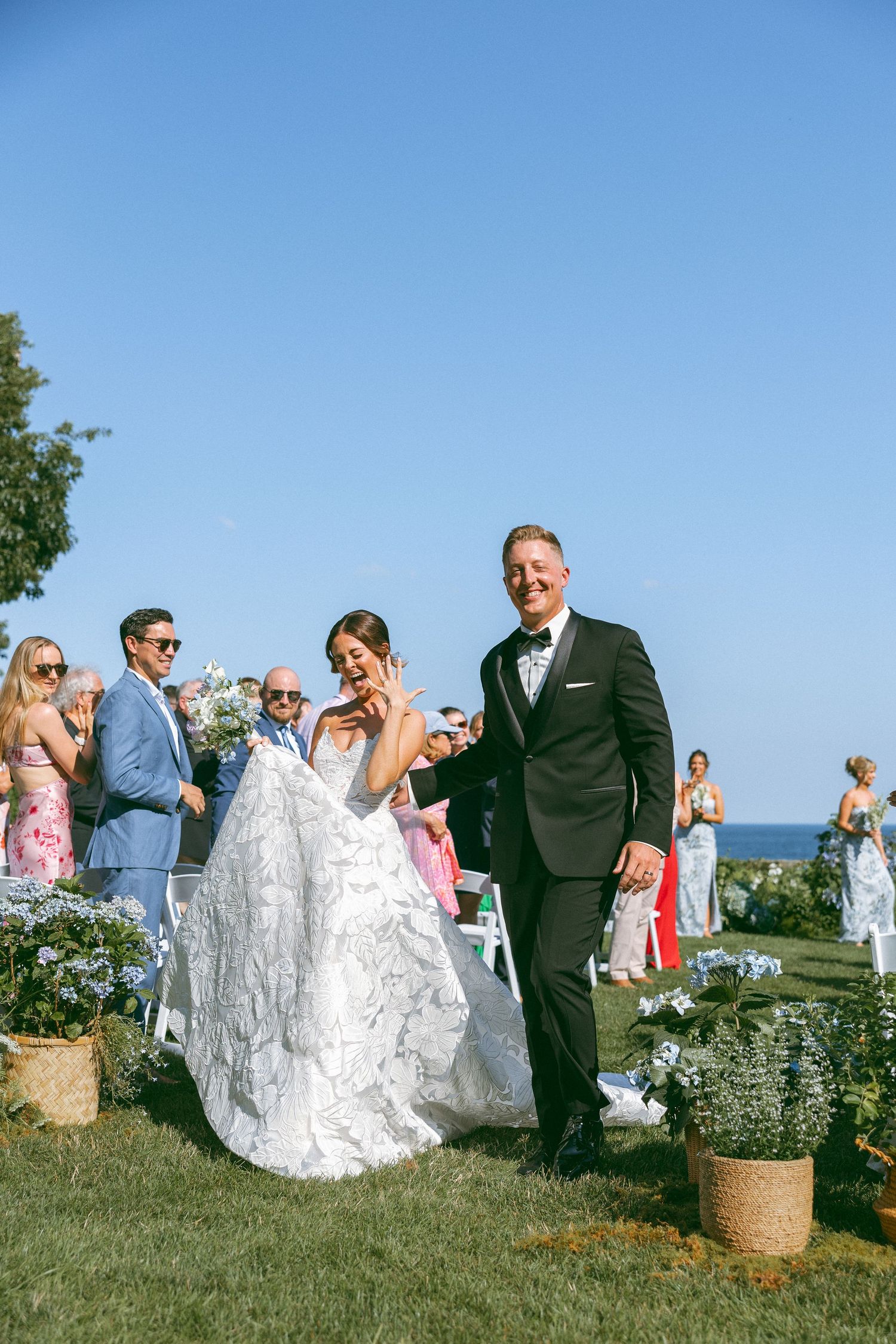 Wedding guests gather on a seaside lawn as a couple walks through in formal attire.