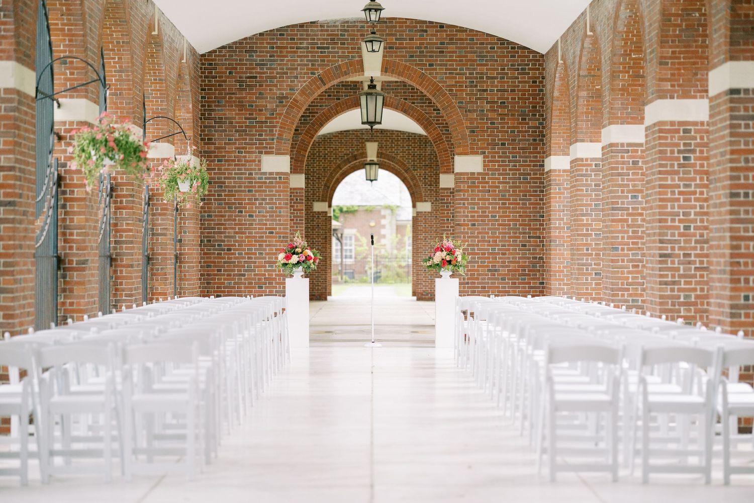 White chairs line a symmetrical brick archway corridor with hanging light fixtures creating an elegant wedding ceremony venue.