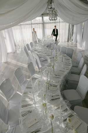 Long elegant banquet tables with white linens and chair covers set up under a draped tent ceiling with chandeliers.