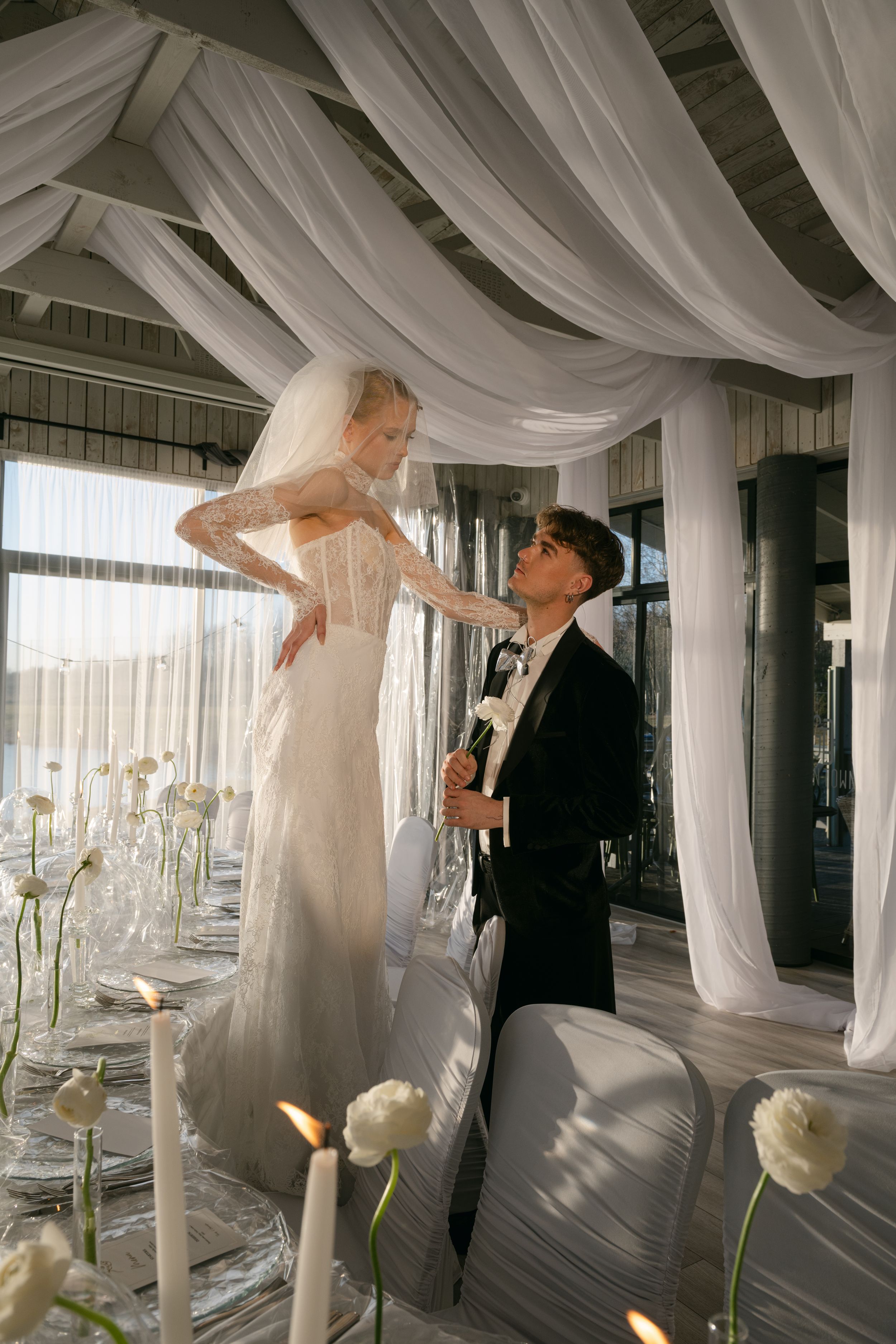 Elegant wedding photoshoot in a light-filled venue with white fabric draping and warm lighting effects.