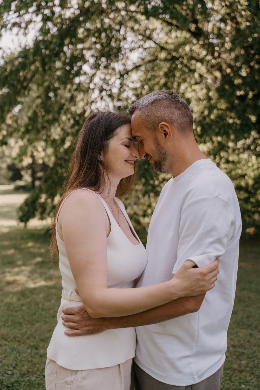 Ein Paar teilt in weißen Oberteilen einen intimen Moment im Freien in einem sonnendurchfluteten Garten. Mama und Papa Moment.