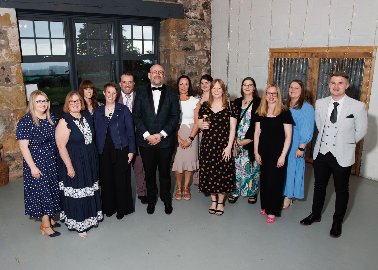 A group photo of formally dressed guests at a wedding celebration in a rustic stone building venue.