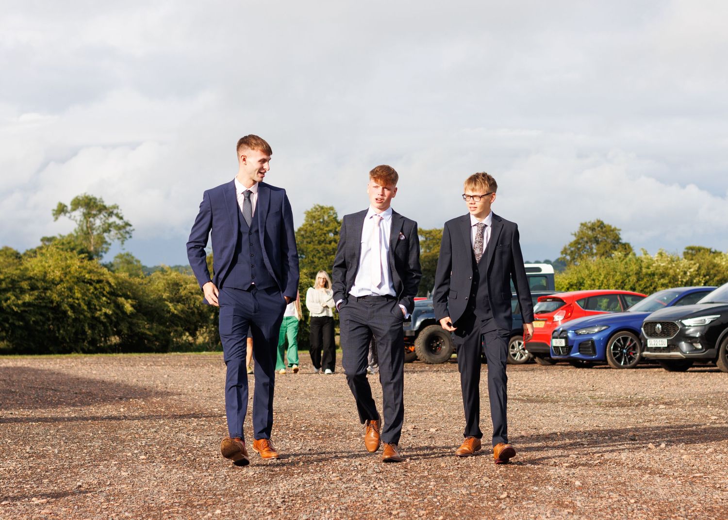 Three groomsmen in stylish suits walk through a parking area on a cloudy day.
