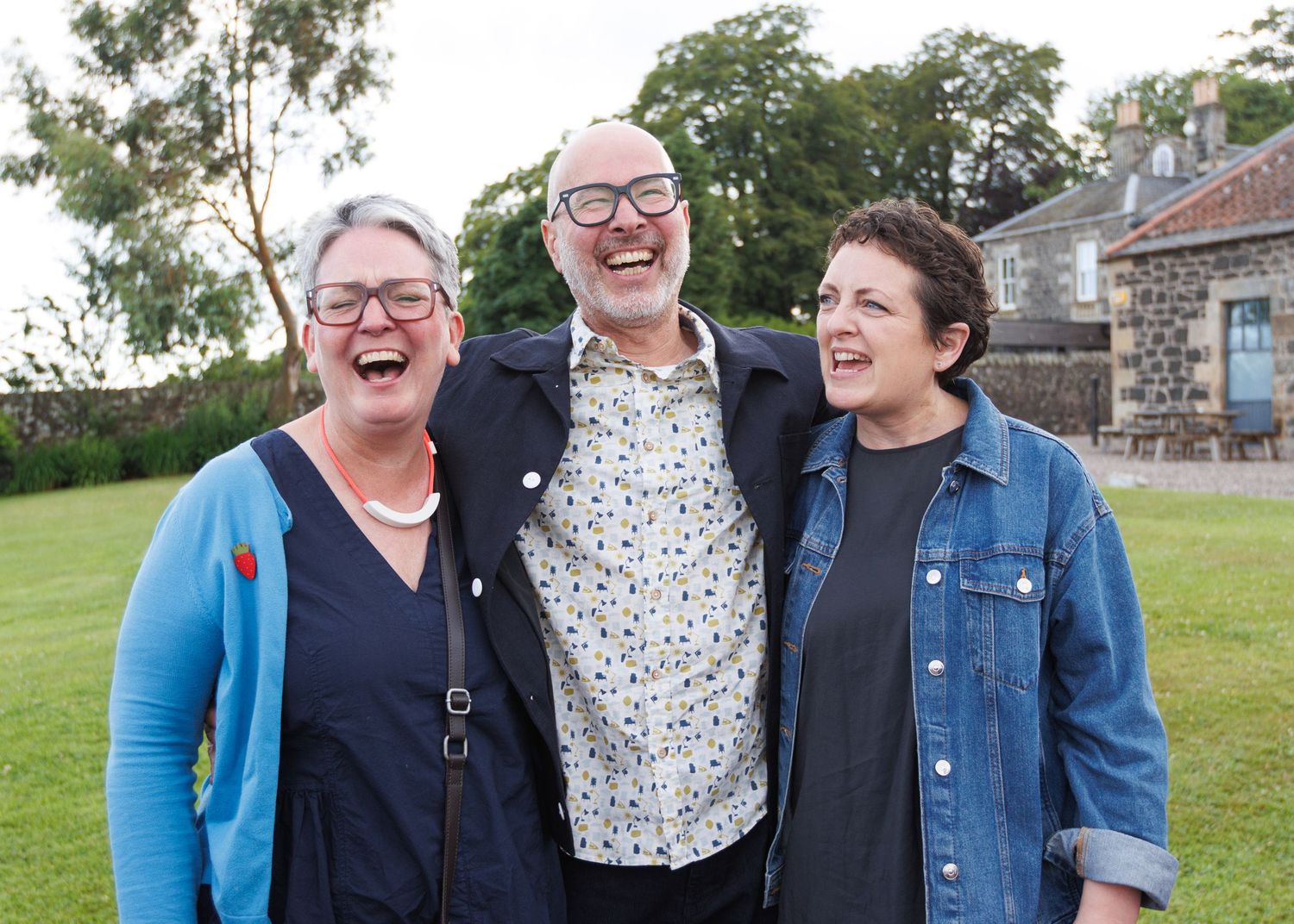 Friends laughing together outdoors on a sunny day wearing casual clothing.