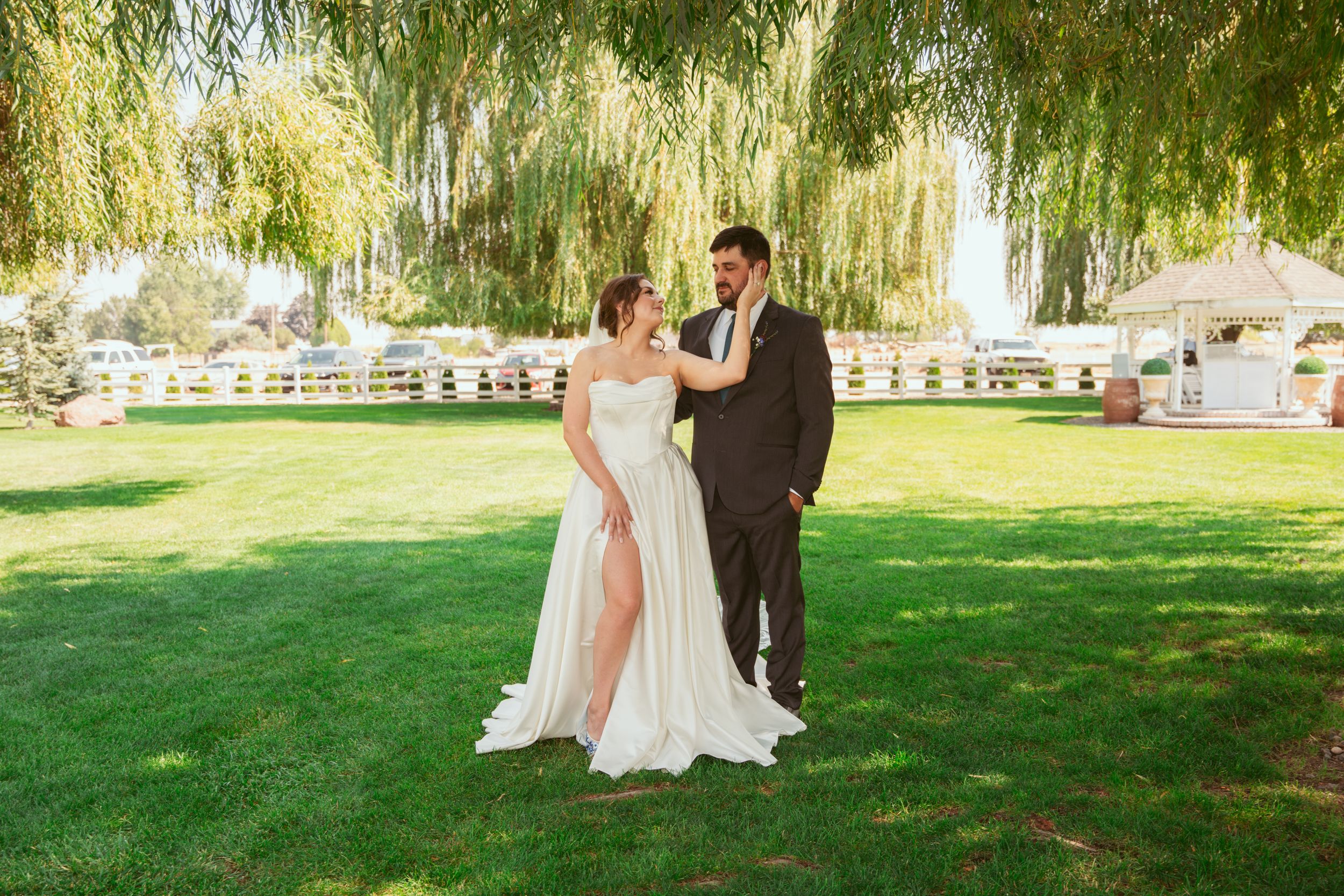 Couple shares a romantic moment on their wedding day under willow trees beside a lake.