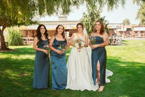 Wedding party in elegant teal blue dresses and white gown pose casually on grassy lawn during outdoor celebration.