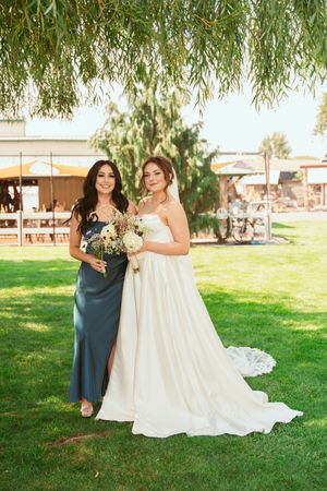 A bride in a flowing white gown poses with her bridesmaid in a teal dress under a willow tree on a sunny day.
