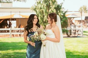A bride and bridesmaid share a joyful moment on a sunny day outdoors, holding a soft pastel floral bouquet.