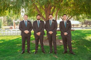 Groomsmen in dark suits stand together on a green lawn under a weeping willow tree at an outdoor wedding venue.