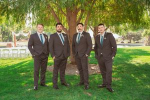 Groomsmen in dark suits standing together under a willow tree on a sunny lawn with white chairs in background.