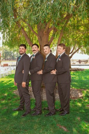 Four groomsmen in matching dark suits pose under a weeping willow tree on a sunny day.