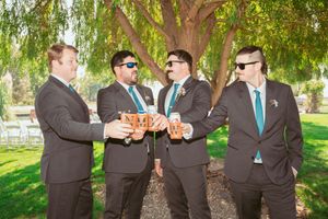 Groomsmen in black suits share drinks and toast while standing under weeping willow trees at an outdoor wedding celebration.
