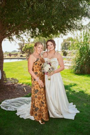 Two women pose outdoors under a tree, one wearing a floral dress and one in a flowing white wedding gown with bouquet.