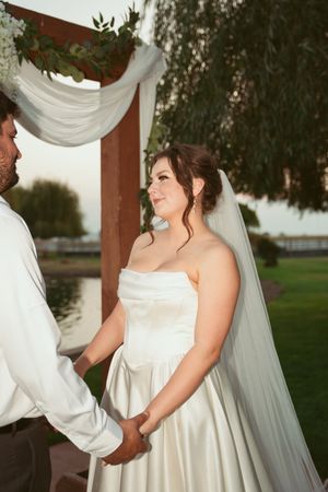 A couple stands together under a white draped arch at an outdoor wedding ceremony by a lake.