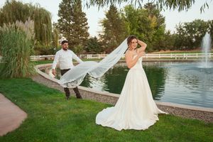 Newlyweds playfully pose by a fountain in a garden setting with bride's veil flowing in the wind.