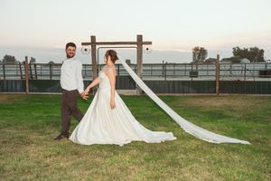 A couple in wedding attire pose together on a grassy lawn at sunset with a wooden cross structure behind them.