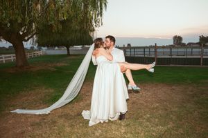 Elegant bride in flowing white gown and groom pose together under a weeping willow tree at dusk.