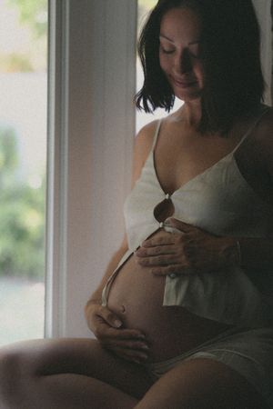 Soft window light illuminates intimate maternity moment in white lingerie.