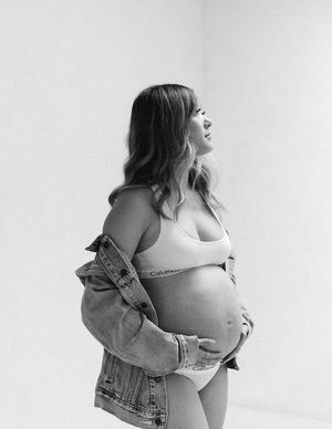 Black and white photo of a pregnant woman in underwear and denim jacket posing against a light background.