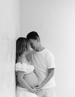 Black and white portrait of a couple sharing an intimate moment while holding a pregnant belly against a white wall.