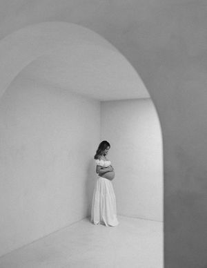 Moody black and white photograph of a figure in a flowing white dress standing in a minimalist arched alcove.