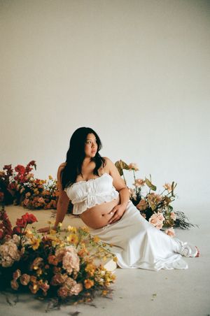 Beautiful maternity photo with white dress among colorful floral arrangements against neutral backdrop.