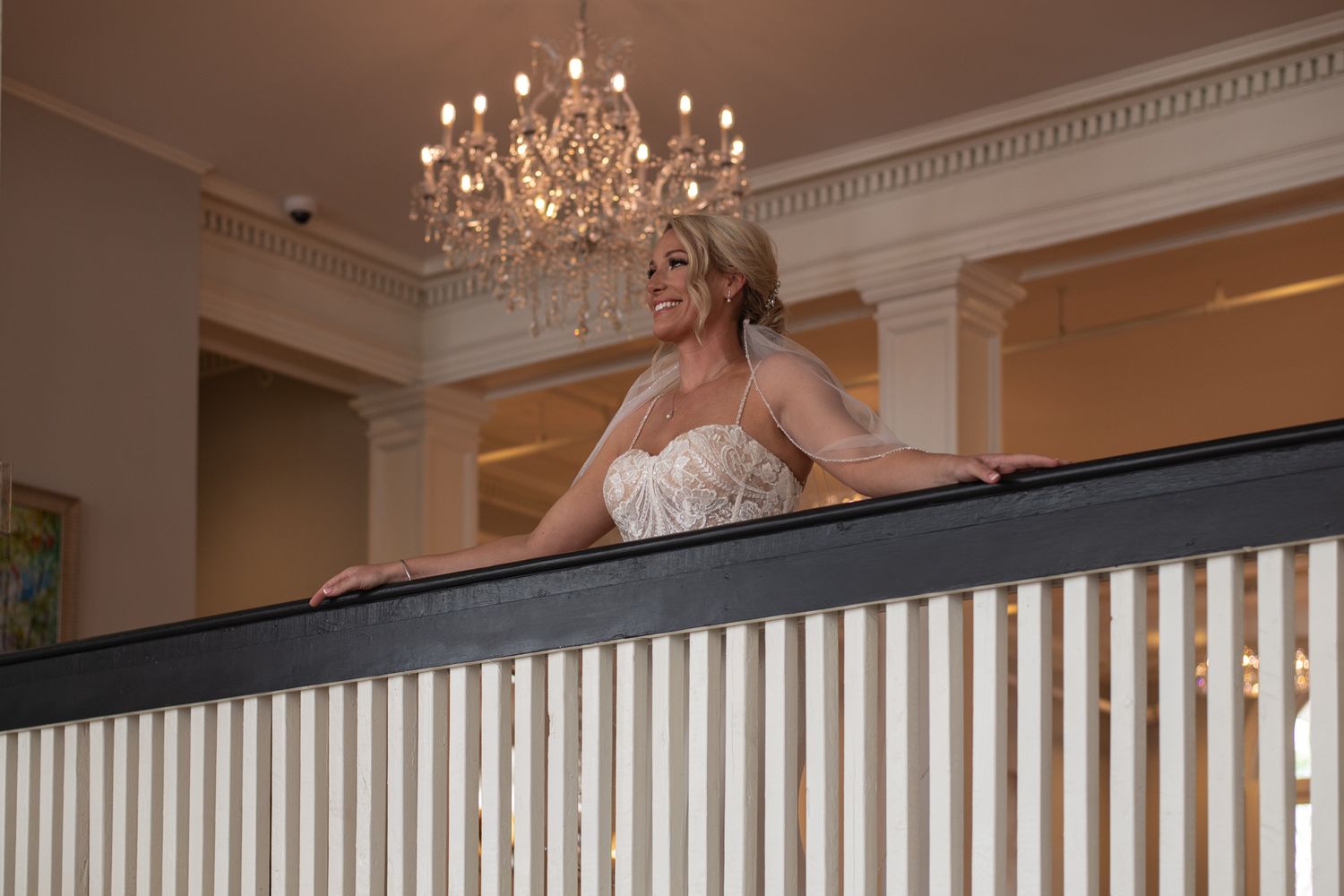 An elegant bride in a white dress is standing on a balcony beneath a crystal chandelier.