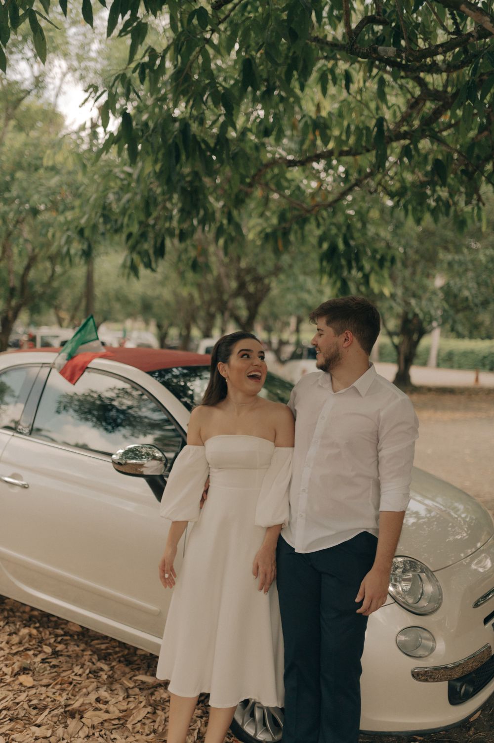 A happy couple posing near a vintage Fiat 500 car under trees.