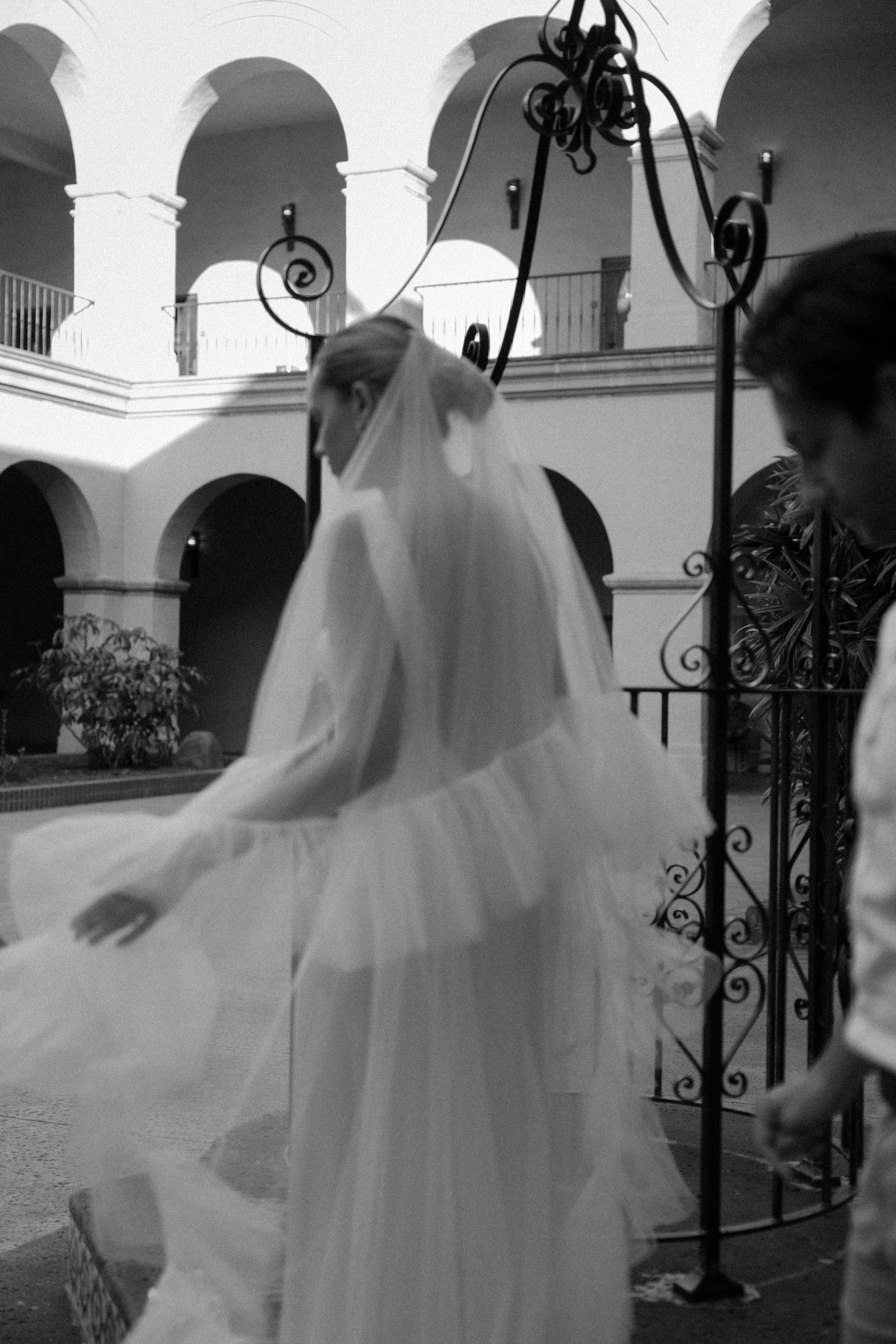 A black and white photo of a bride in flowing white dress and veil walking through an arched courtyard.