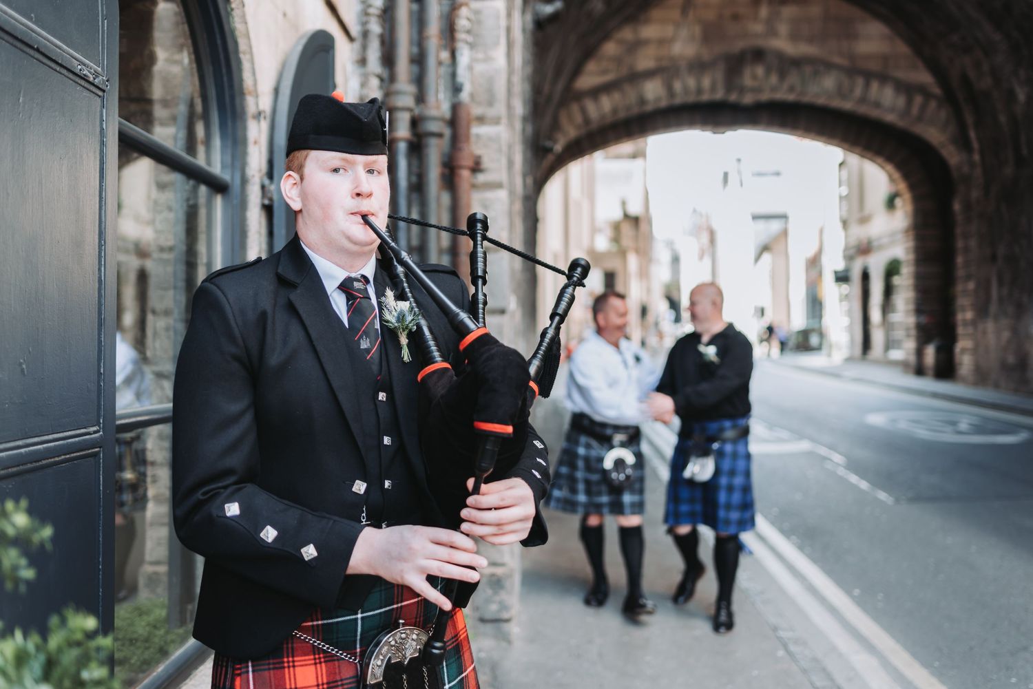 Traditional Scottish bagpiper performing in traditional dress under a stone archway on a city street.