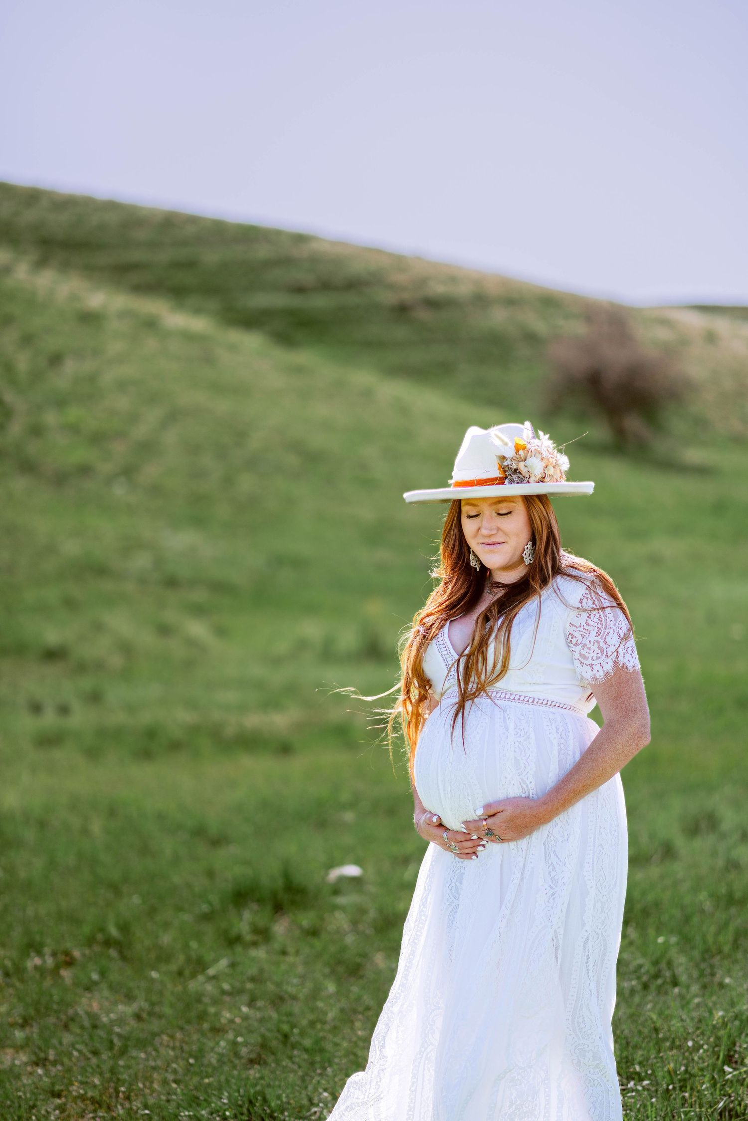 Maternity photo in flowing white dress on grassy hillside with floral decorated hat.