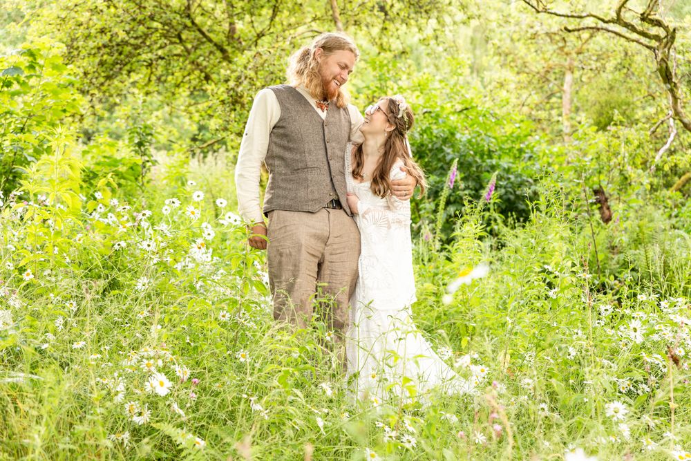 A wedding portrait of a bride and groom posing cuddled up to one another amongst wildflowers in a woodland glade.