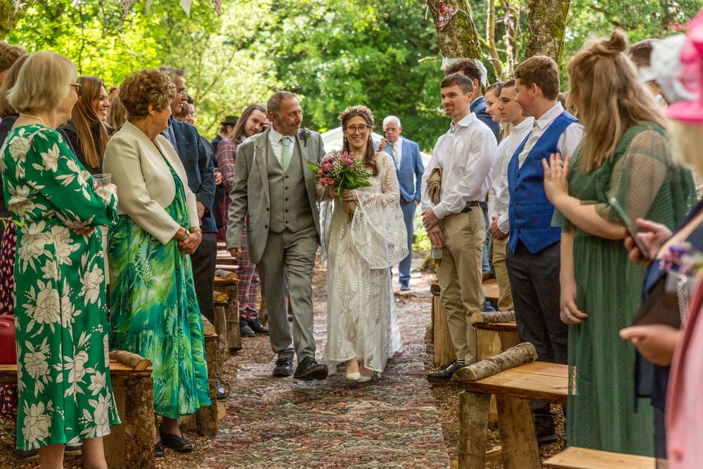 A bride walks down the aisle with her father in a woodland glade at her outdoor pagan wedding.
