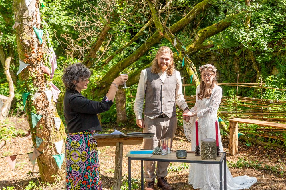 Handfasting photography. A couple stand holding hands in a woodland glade as a celebrant conducts their handfasting ceremony.