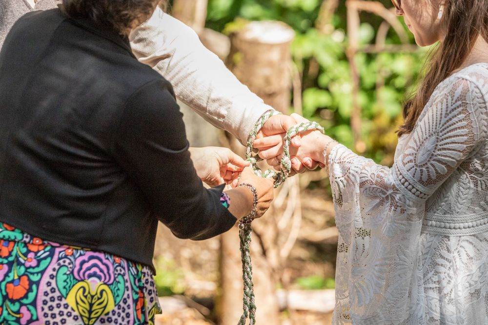 A celebrant ties a couple's hands with a woven cord during an outdoor handfasting ceremony.