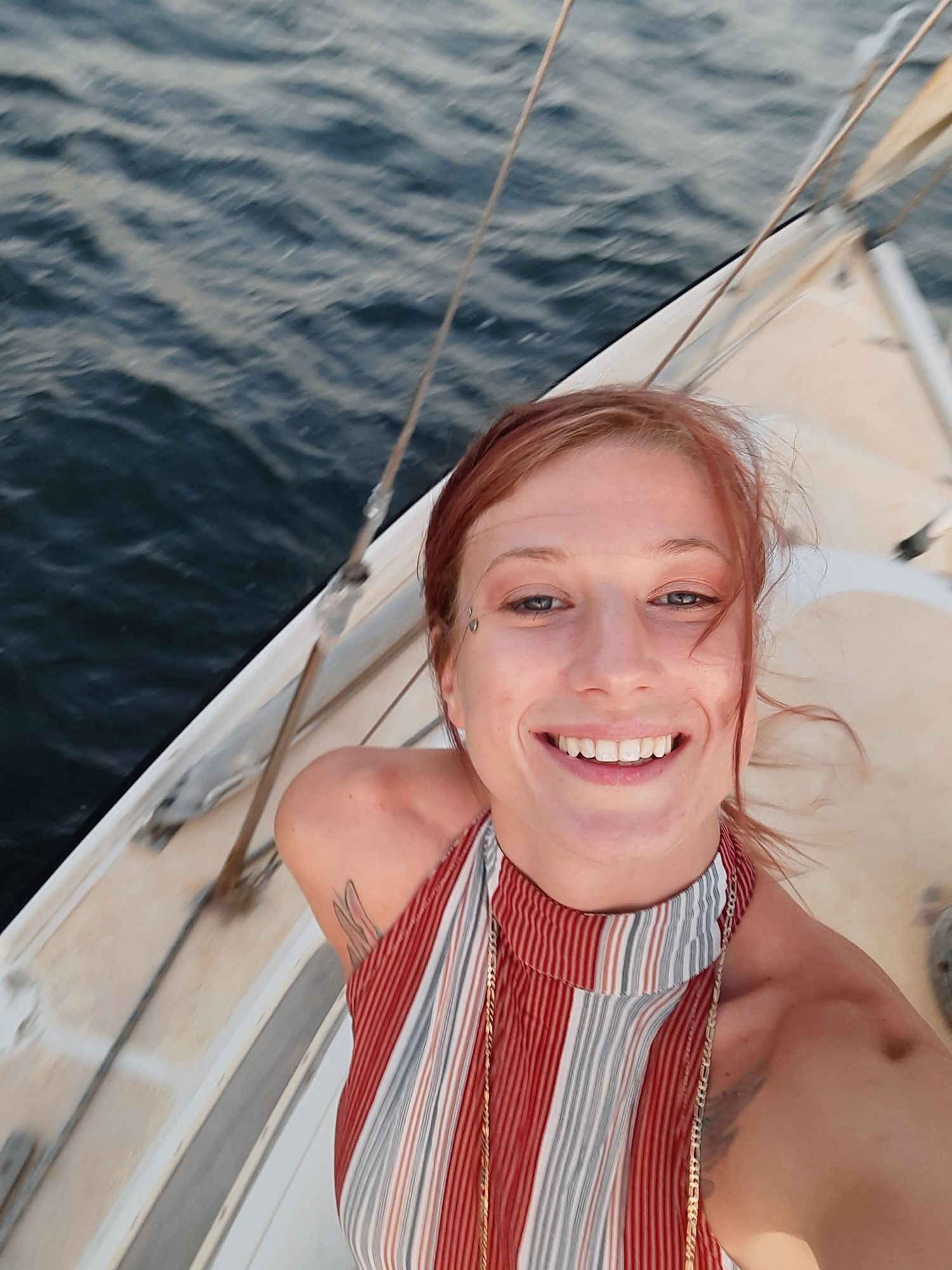 A person takes a selfie on a sailboat against blue rippling water on a sunny day.
