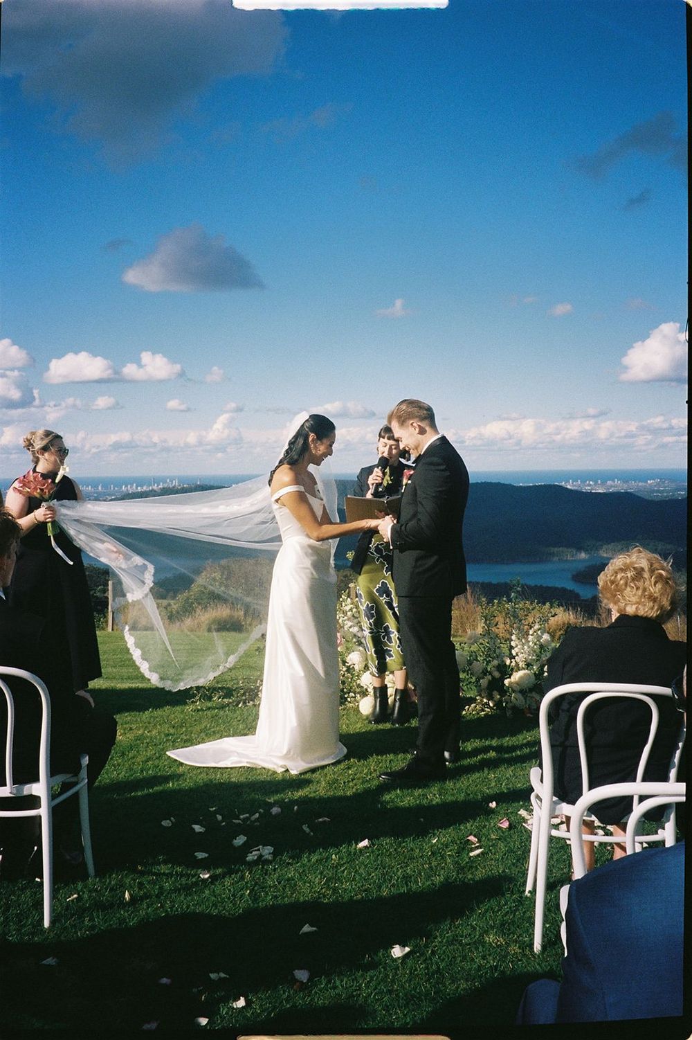 An outdoor wedding ceremony set against scenic mountain views with white chairs and flowing veils catching the breeze.