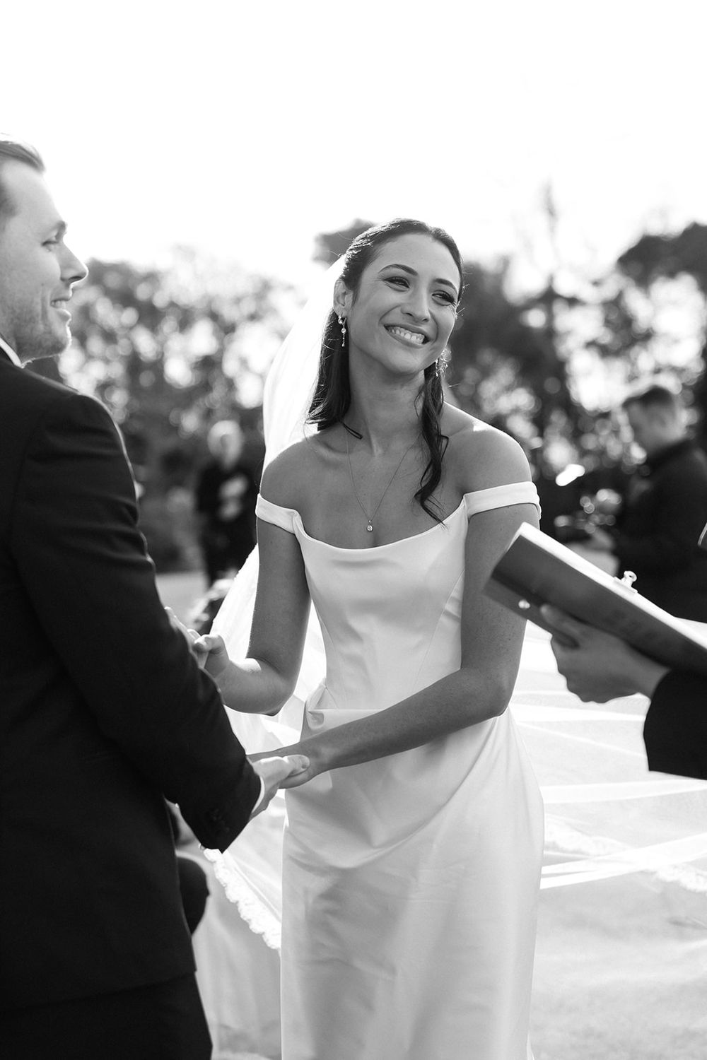 Bride in an elegant off-shoulder white gown twirls joyfully during outdoor wedding celebration.
