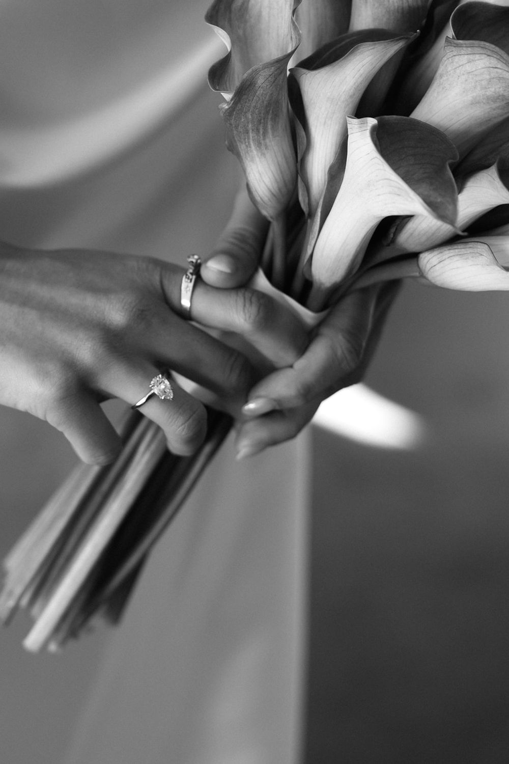 Black and white close-up of hands holding a bouquet of calla lilies with diamond rings displayed on fingers.