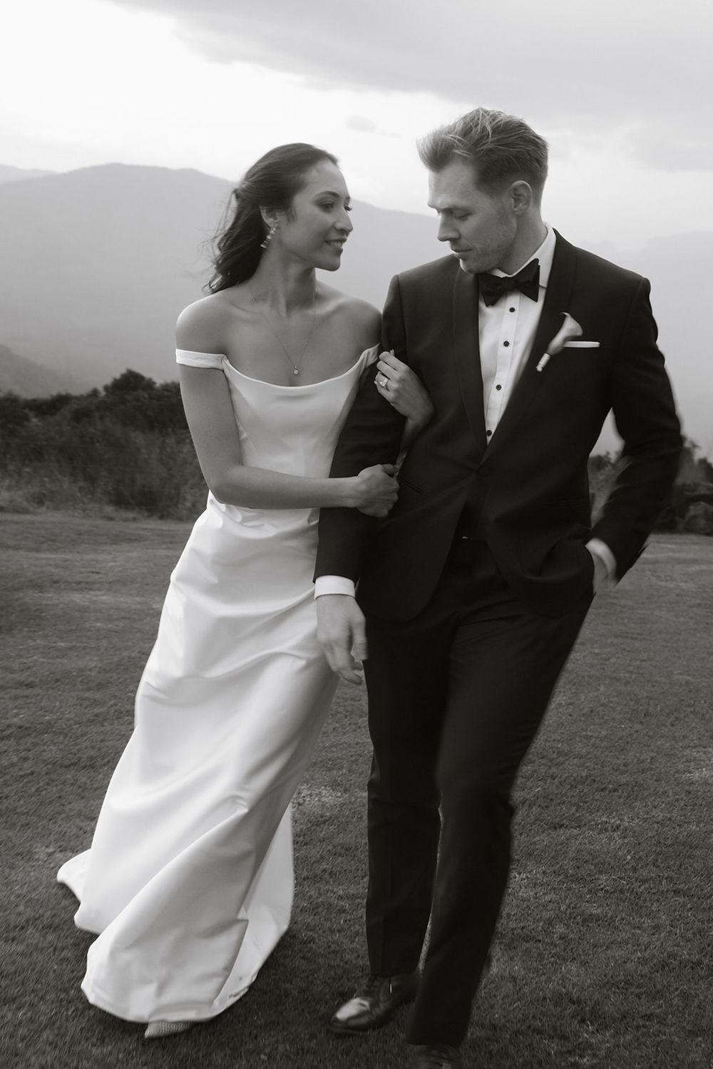 Black and white wedding photo of newlyweds walking together on a hilltop with mountains in background.