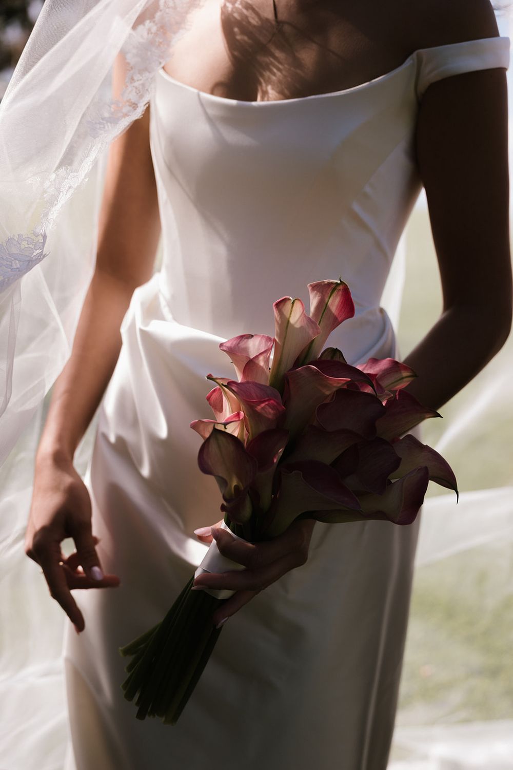 A bride in an elegant white dress holds a bouquet of deep burgundy calla lilies against soft sunlit background.