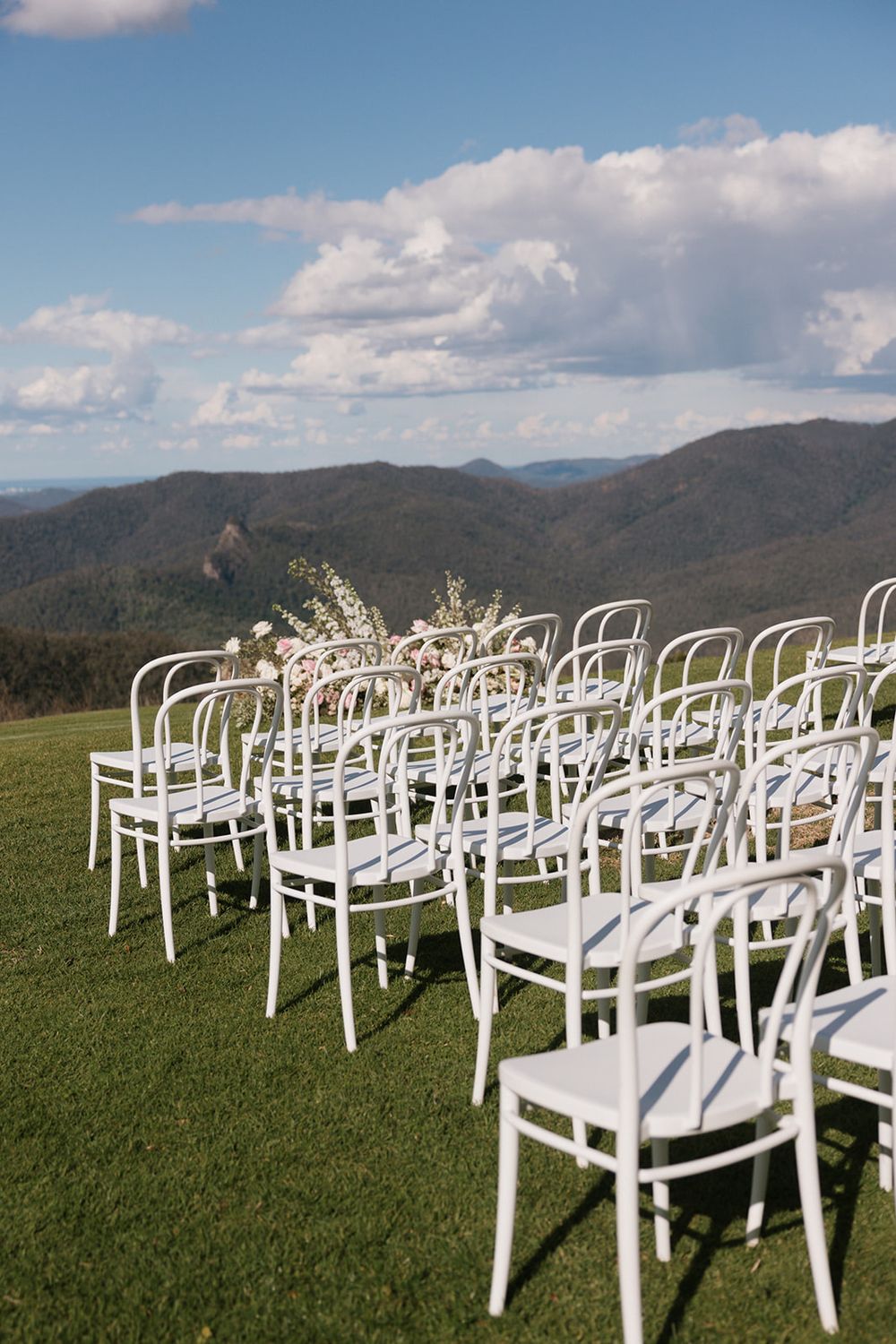 White chairs arranged in rows for an outdoor wedding ceremony against a scenic mountain backdrop on a grassy hilltop.