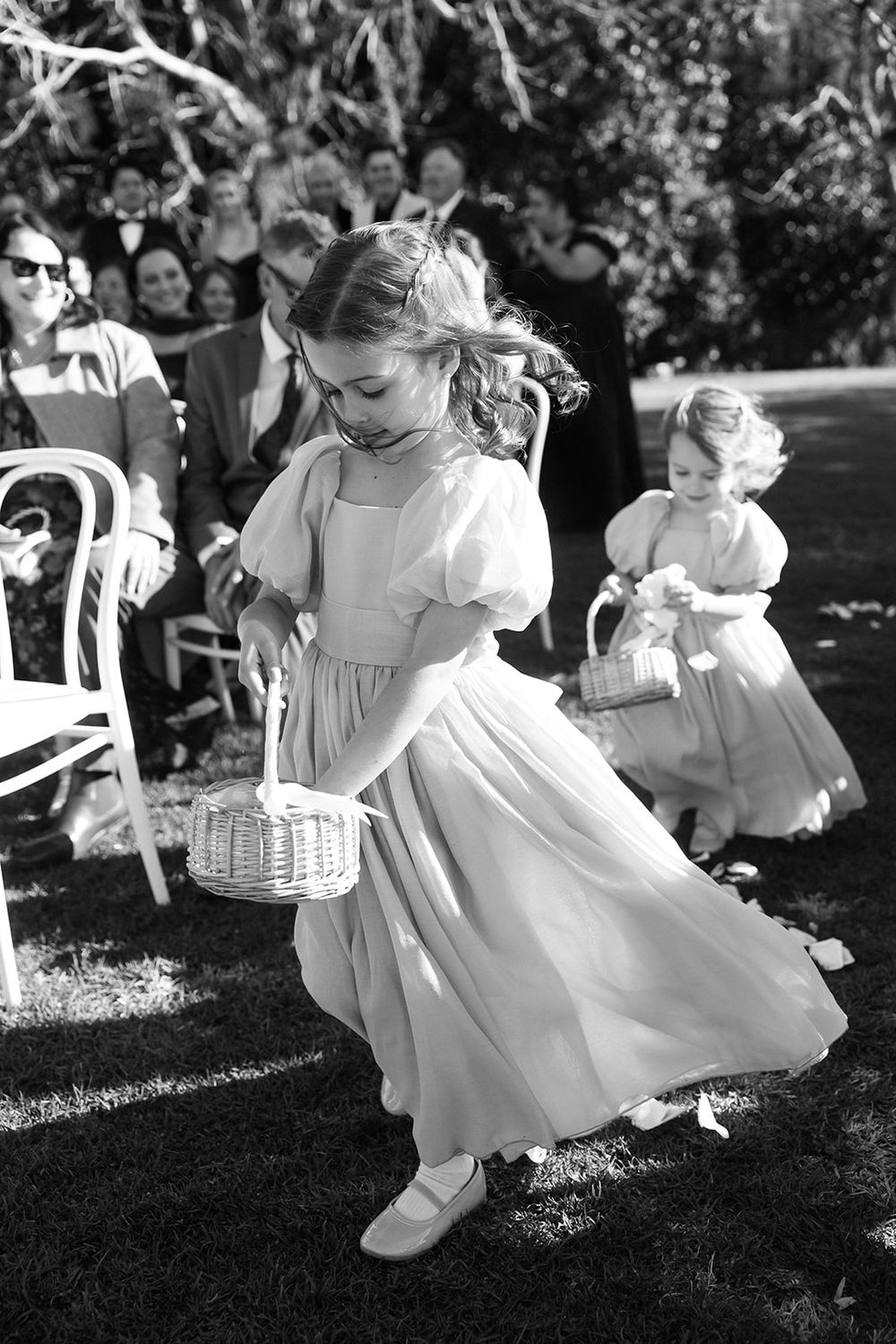 Small children in white dresses and clothing play and dance together at an outdoor event on a sunny day.