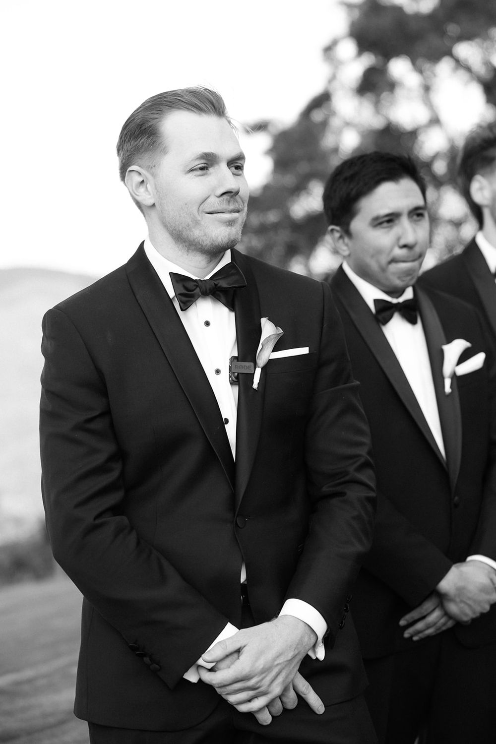 Groomsmen in black tuxedos and bow ties share candid laughs during outdoor wedding photo session.