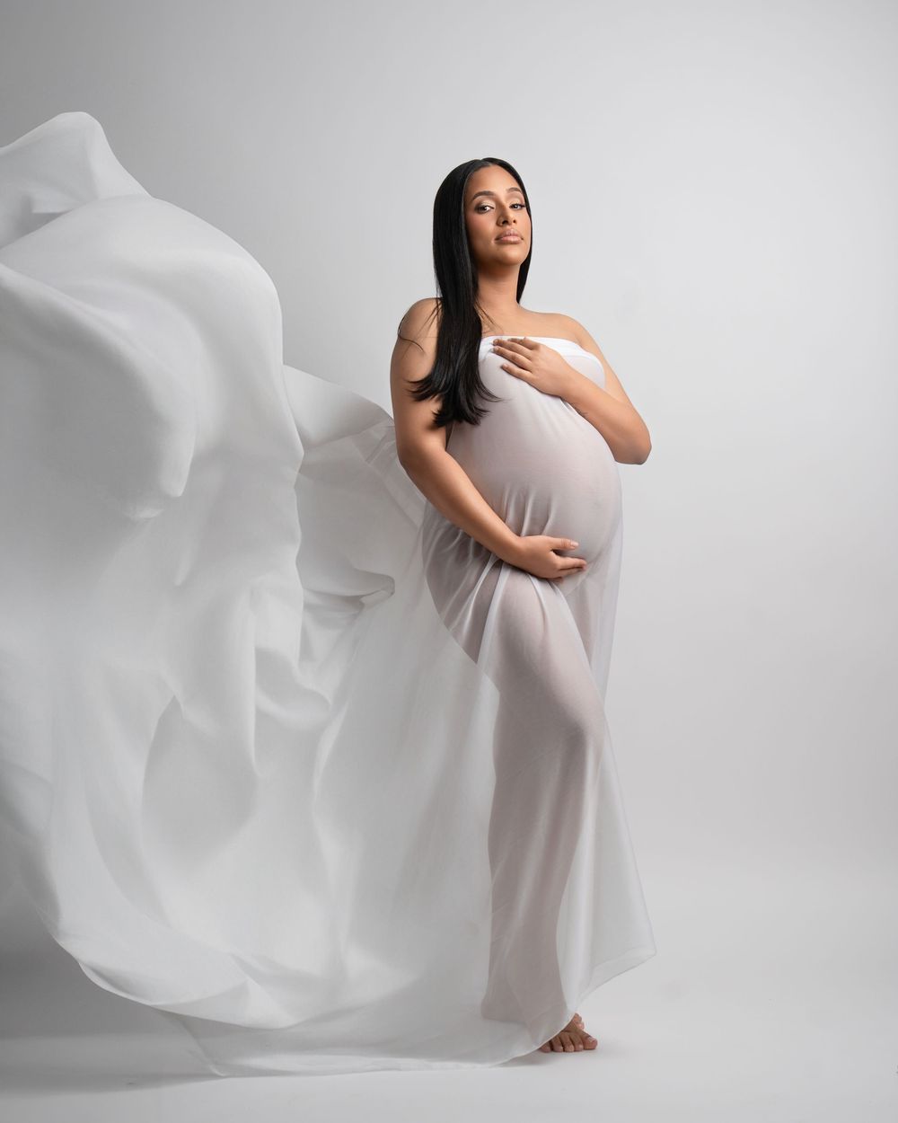 Minimalist maternity portrait with flowing white fabric against light gray background.