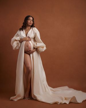 Dramatic maternity portrait in flowing cream robe with long sleeves against terracotta backdrop.