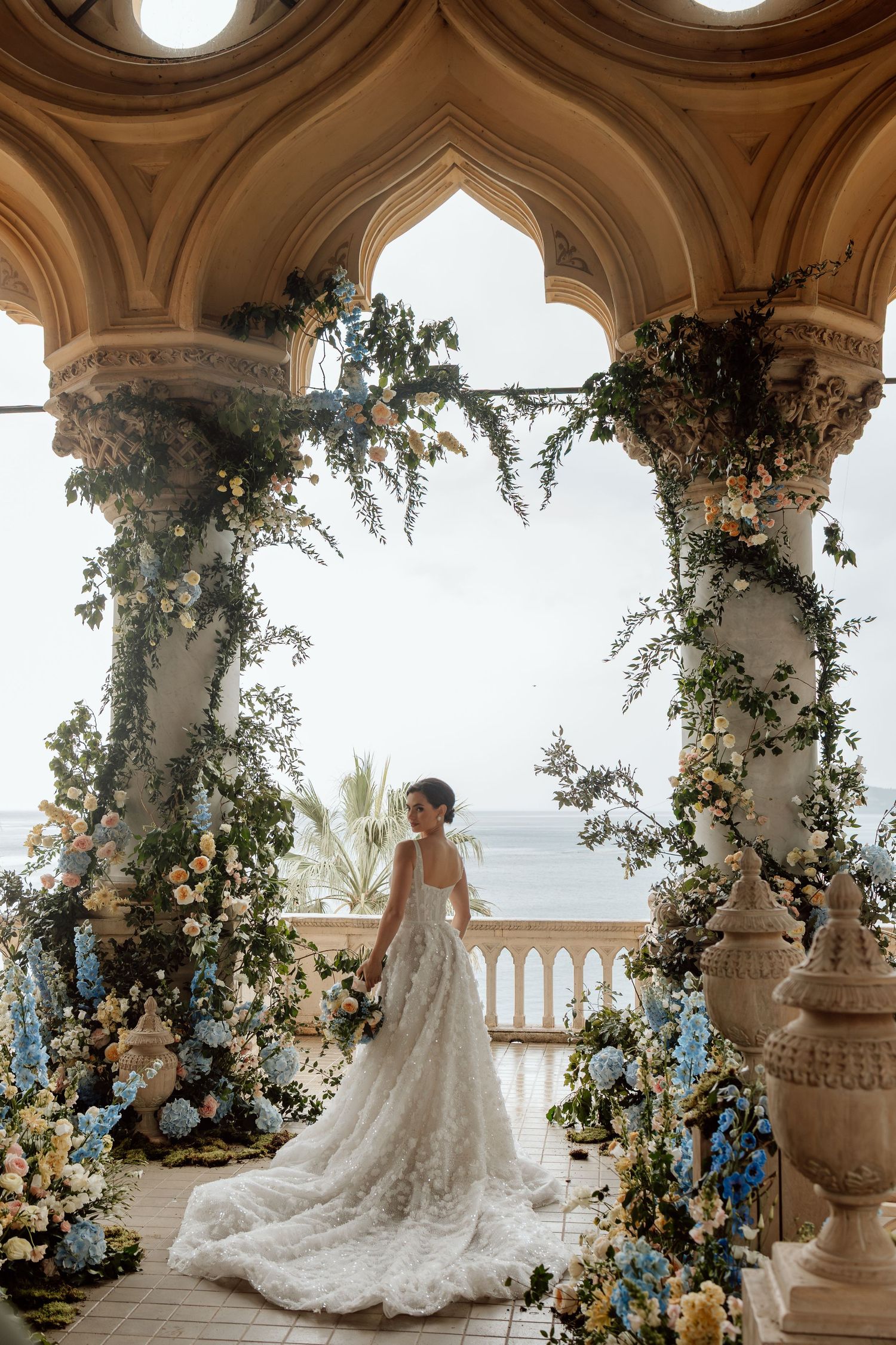 Elegant Italy wedding scene with floral archways and ocean view from ornate balcony featuring white lace gown with flowing train.