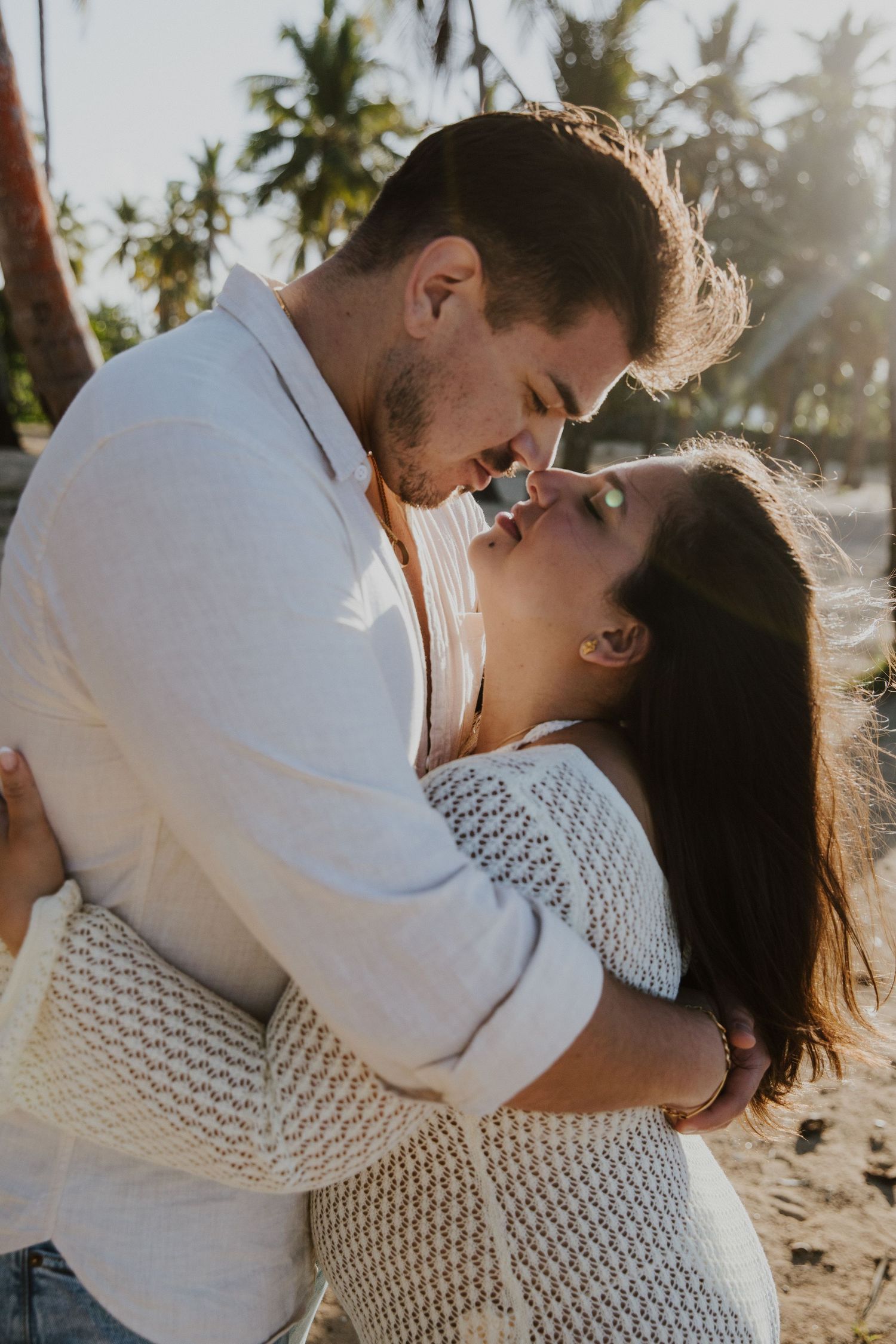 A tender embrace between a couple on a sunny beach with palm trees in the distance.