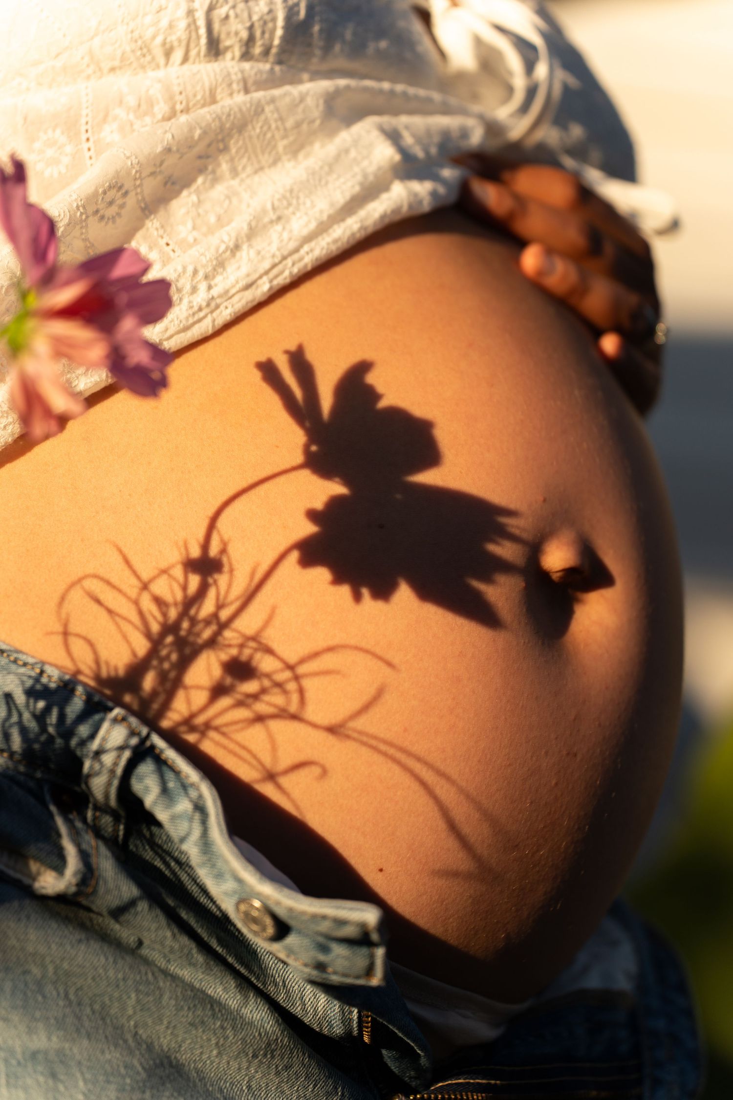 Pregnant woman's bare belly with flower shadow cast by golden sunlight, wearing denim jeans and white top. Maternity floral session