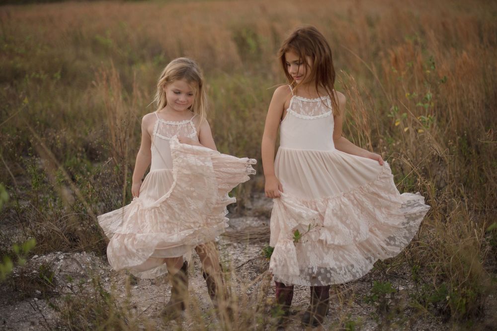 Two children in matching white lace dresses twirling in a grassy field at sunset.